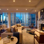 Large open plan apartment with living room area with fireplace, dining table, and windows on two sides with city views.