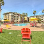 Exterior of lawn, pool deck and pool surrounded by palm trees and 4-story apartment complex