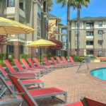 Exterior of pool with brick pool deck rimmed with lounge chairs with 4-story apartment buildings in background