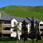 Exterior of a 3 story building with balconies, pitched roof, and a green hill in the background.