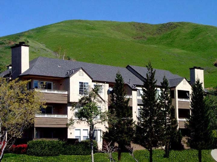 Exterior of a 3 story building with balconies, pitched roof, and a green hill in the background.