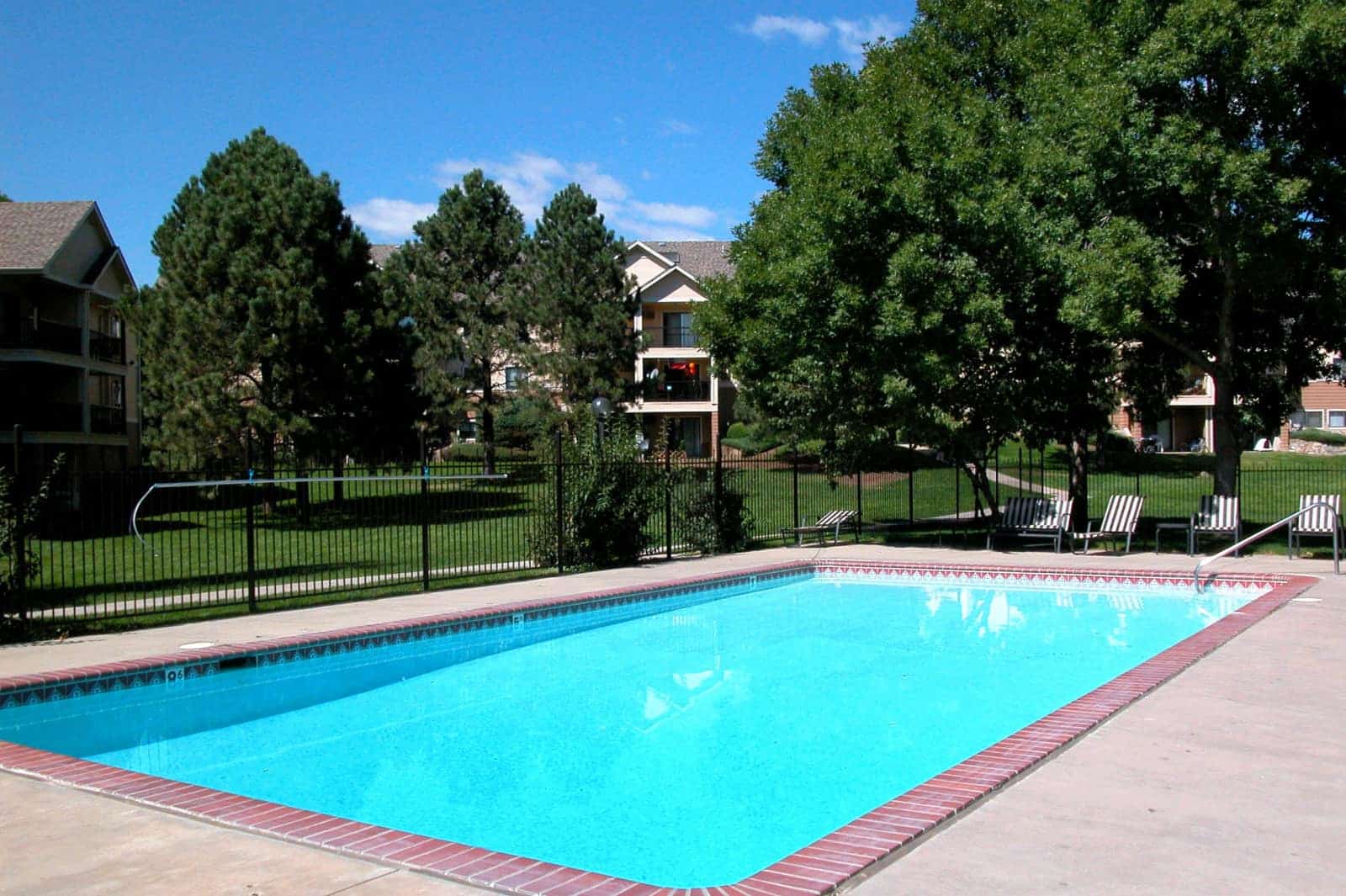 Outdoor pool surrounded by lawn and trees and apartment buildings.