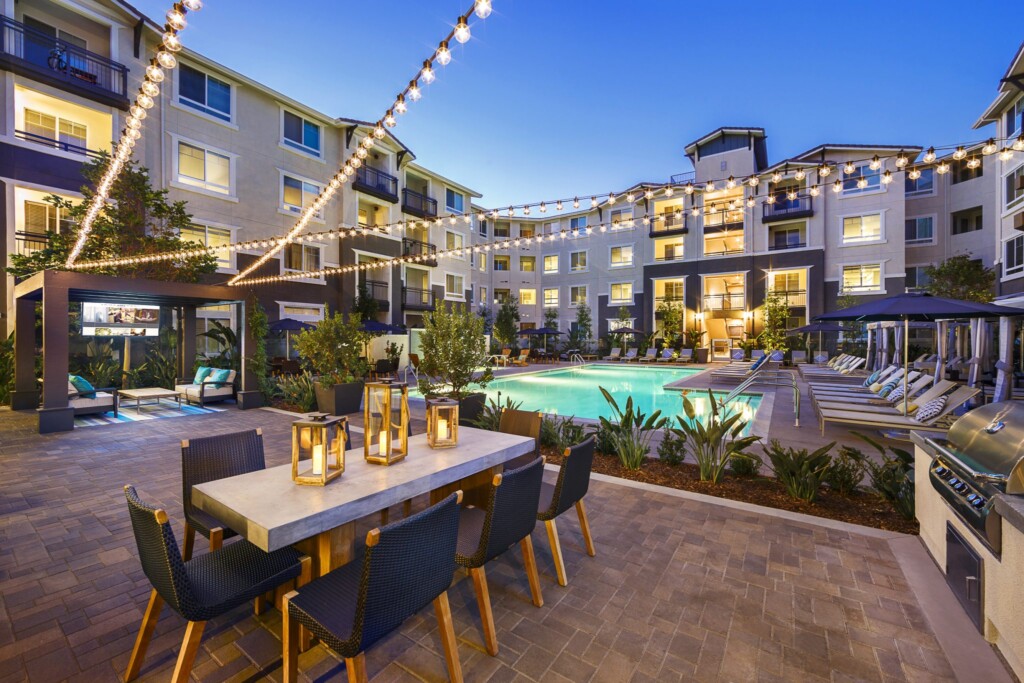Exterior of apartment complex courtyard with barbeque, dining table and cafe lights overhead with pool in background