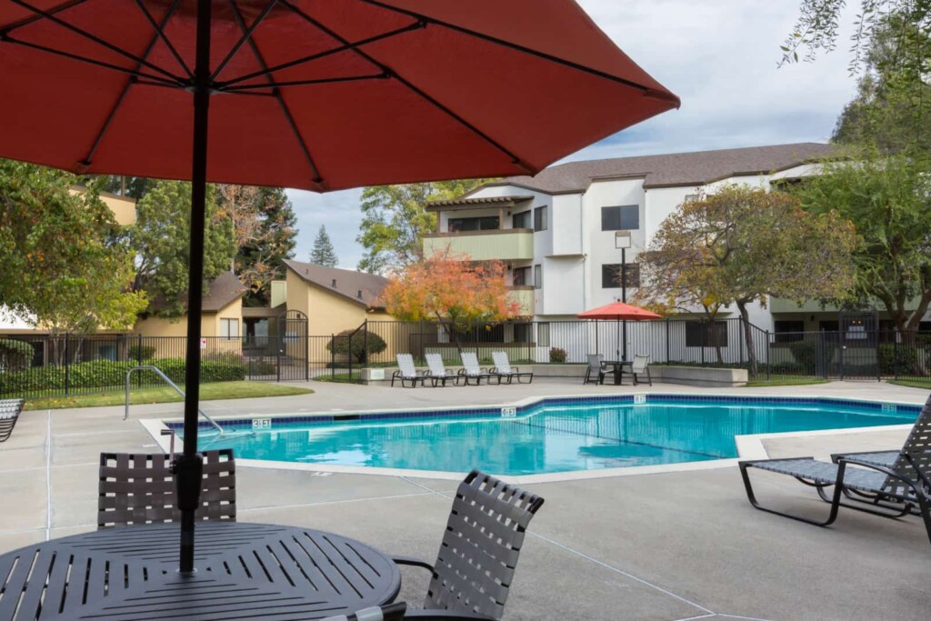 Exterior of pool with table and umbrella in foreground with 3-story apartment buildings in back ground.