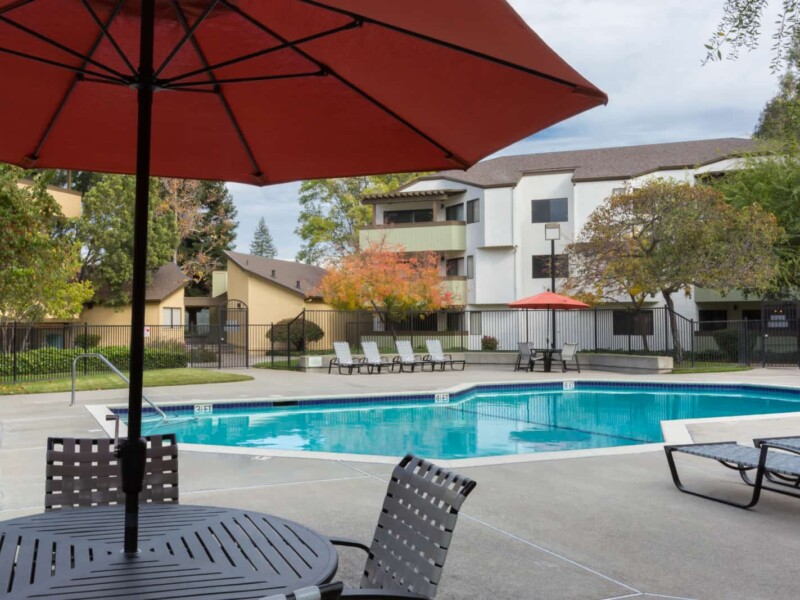 Exterior of pool with table and umbrella in foreground with 3-story apartment buildings in back ground.
