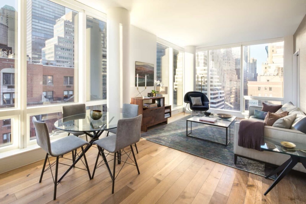 Modern apartment living room with windows on two side, dining table, and city views.