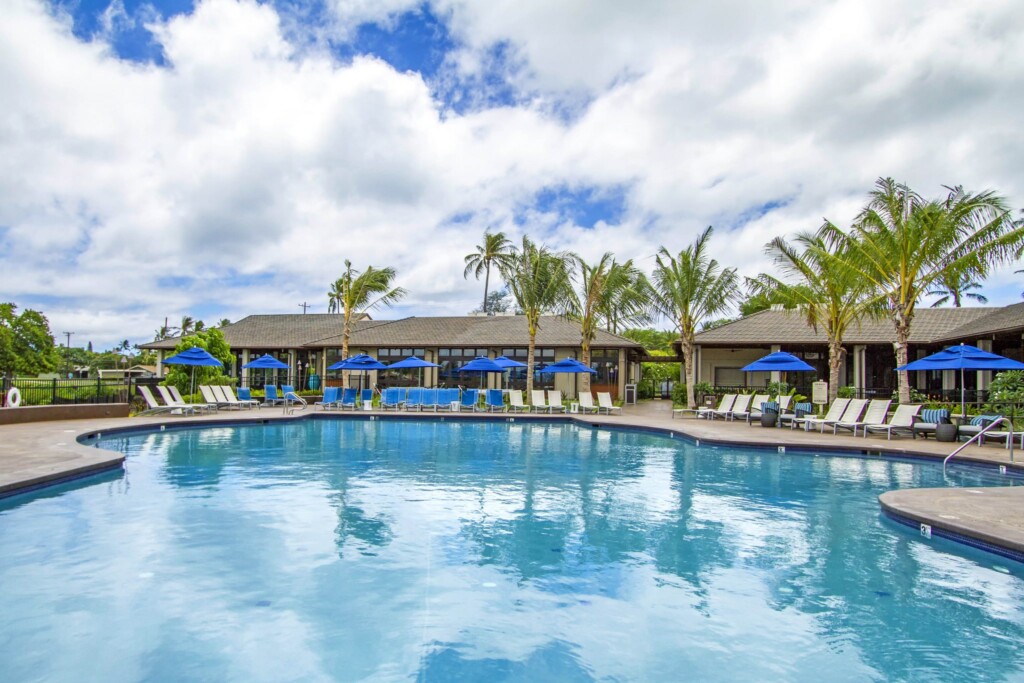 Exterior of swimming pool in foreground surrounded by lounge chairs and single-story buildings.