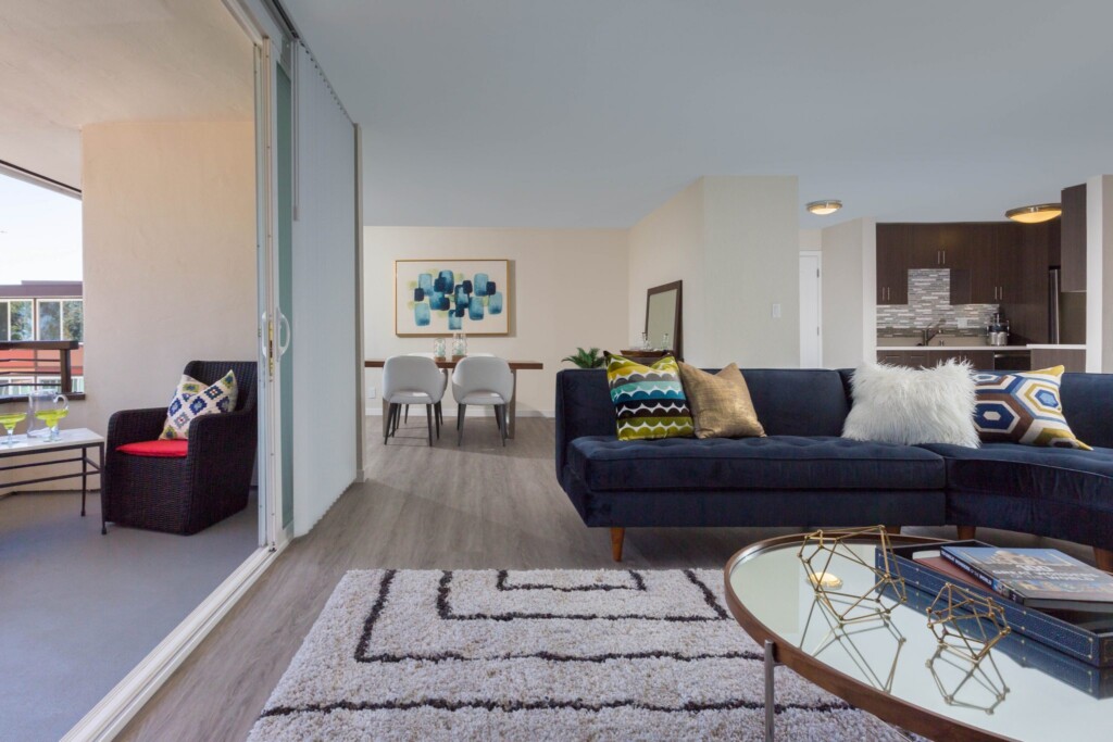 Open floor plan apartment interior of living room with dining area and kitchen in background and patio deck to the side.