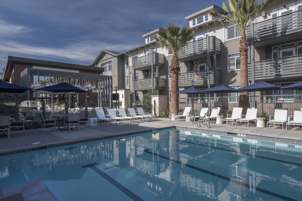 Exterior of pool with lounge chairs and umbrellas with 3-story apartment building with balconies in the background