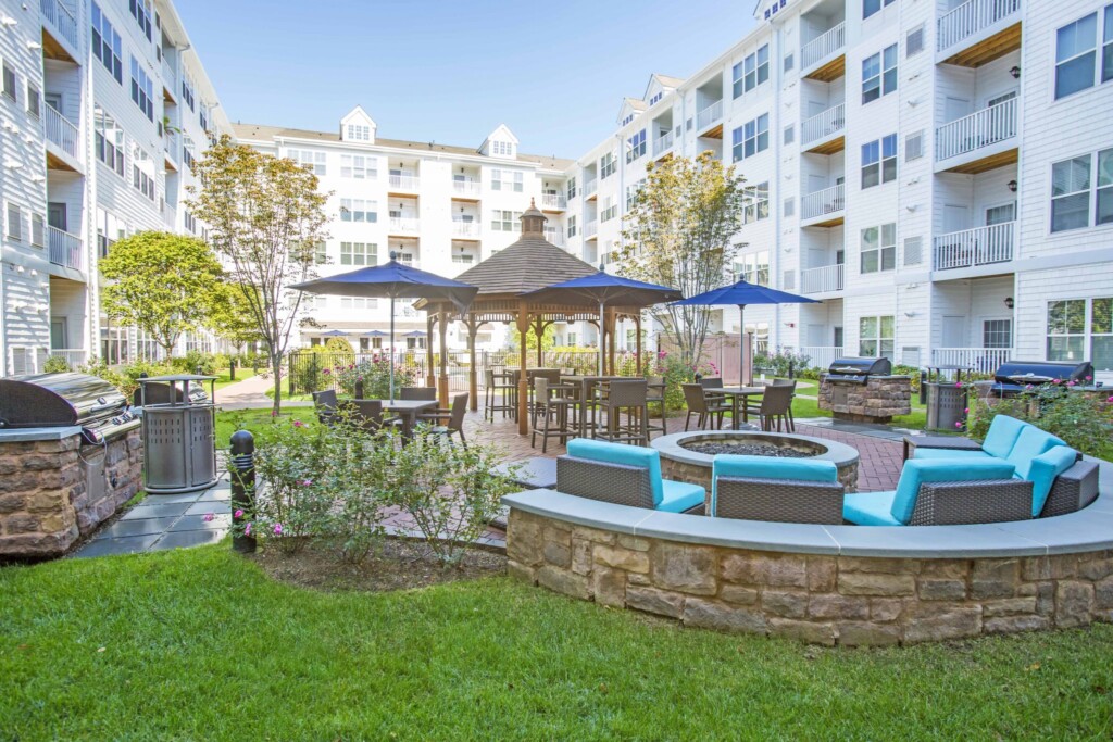 Exterior of courtyard between buildings with grass, seating around a fire pit, barbeques, and table seating.